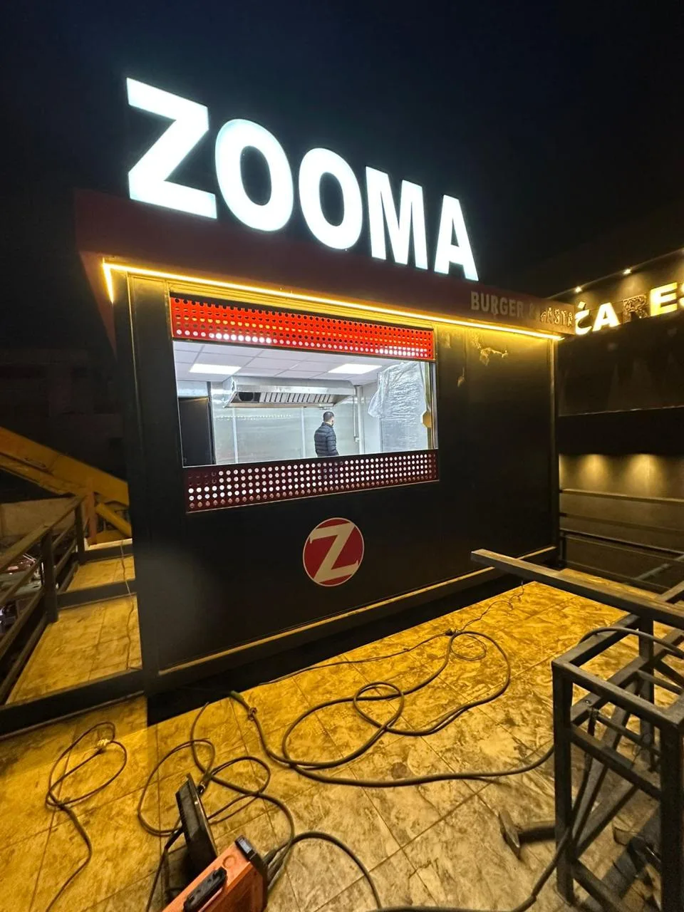 كشك طعام بلافتة ZOOMA مضاءة ليلاً وشباك تقديم بإضاءة حمراء