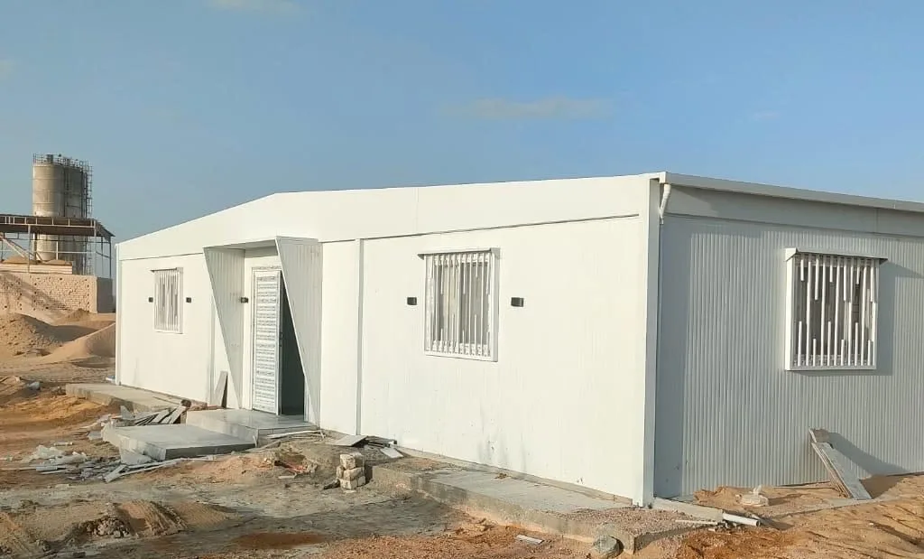 عمال يركّبون مدخل مجمع كرفانات أبيض بنوافذ بحماية حديد في موقع عمل صحراوي