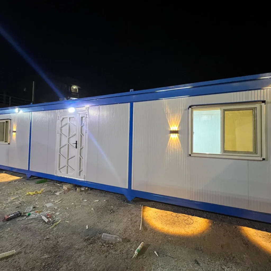 كرفان سكني طويل أبيض بإطار أزرق وباب مزدوج مزخرف ونوافذ بإضاءة جدارية ليلاً