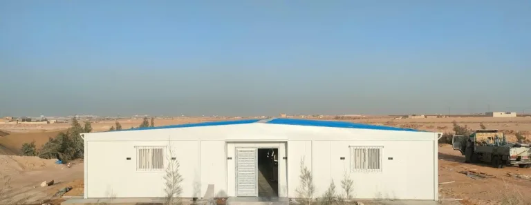 مبنى كرفانات أبيض جاهز بسقف أزرق ومدخل في المنتصف وسط أرض صحراوية