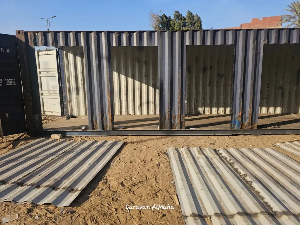 فتحات واسعة مقصوصة في جانب حاوية شحن تمهيداً لتحويلها، مع ألواح الصاج المقصوصة أمامها