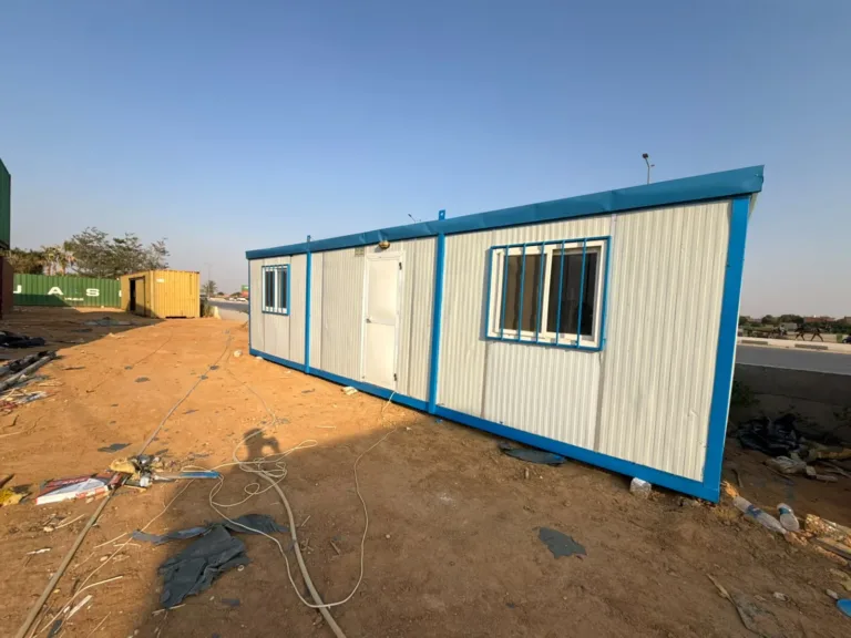 كرفان مستعمل بعد التجديد بألواح بيضاء وإطار أزرق ونافذتين بحماية حديد في ساحة المصنع