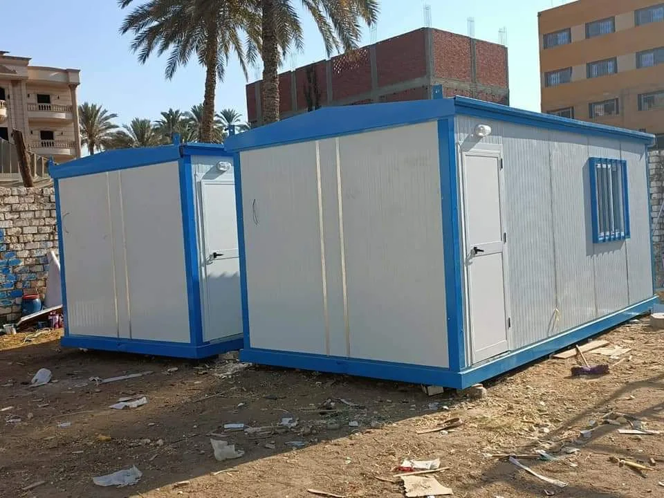 كرفانان من الساندوتش بانل الأبيض بإطار أزرق على أرض ترابية بجوار مبانٍ ونخيل